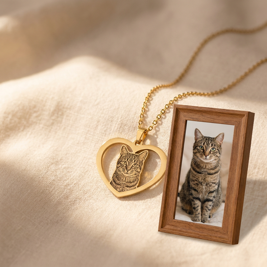 Titanium gold Cat Memorial Heart Photo Necklace with engraved cat portrait pendant on warm cream linen beside a wooden picture frame with a tabby cat photo, cat memorial keepsake by Dazzle Wears