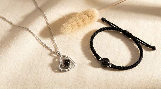 Silver photo necklace and black braided photo bracelet side by side on warm cream linen surface with dried pampas grass