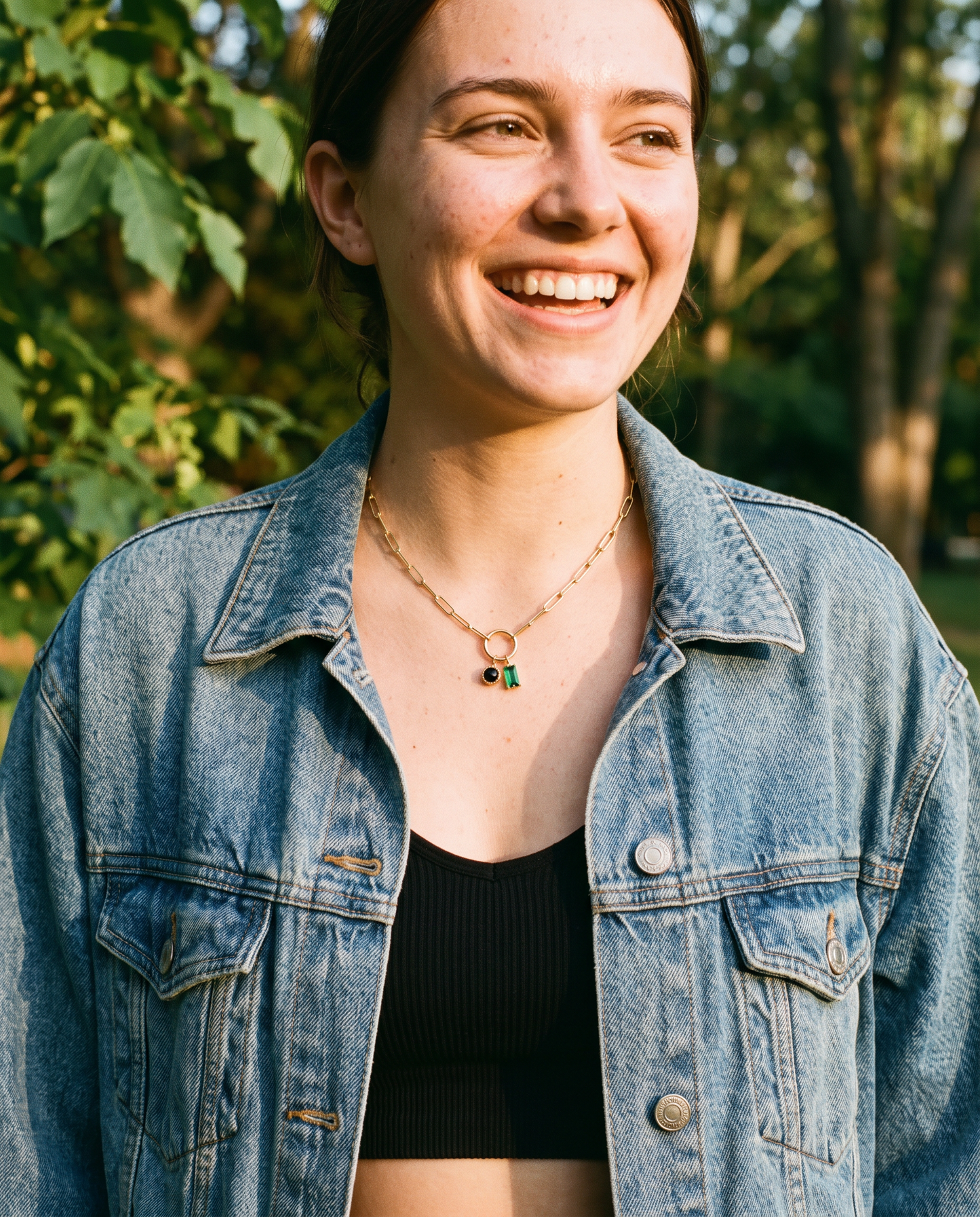 Young woman wearing personalized birthstone charm necklace, paperclip chain, denim jacket outdoors